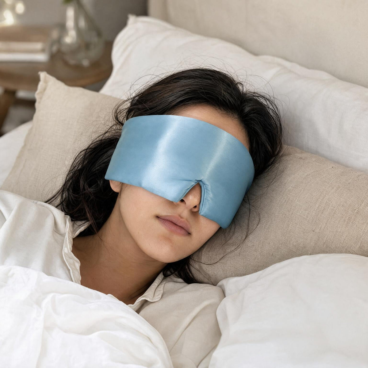 Overhead view of a woman asleep wearing the Lunia Reve mulberry silk sleep mask in Sky Blue, head resting on a linen pillow