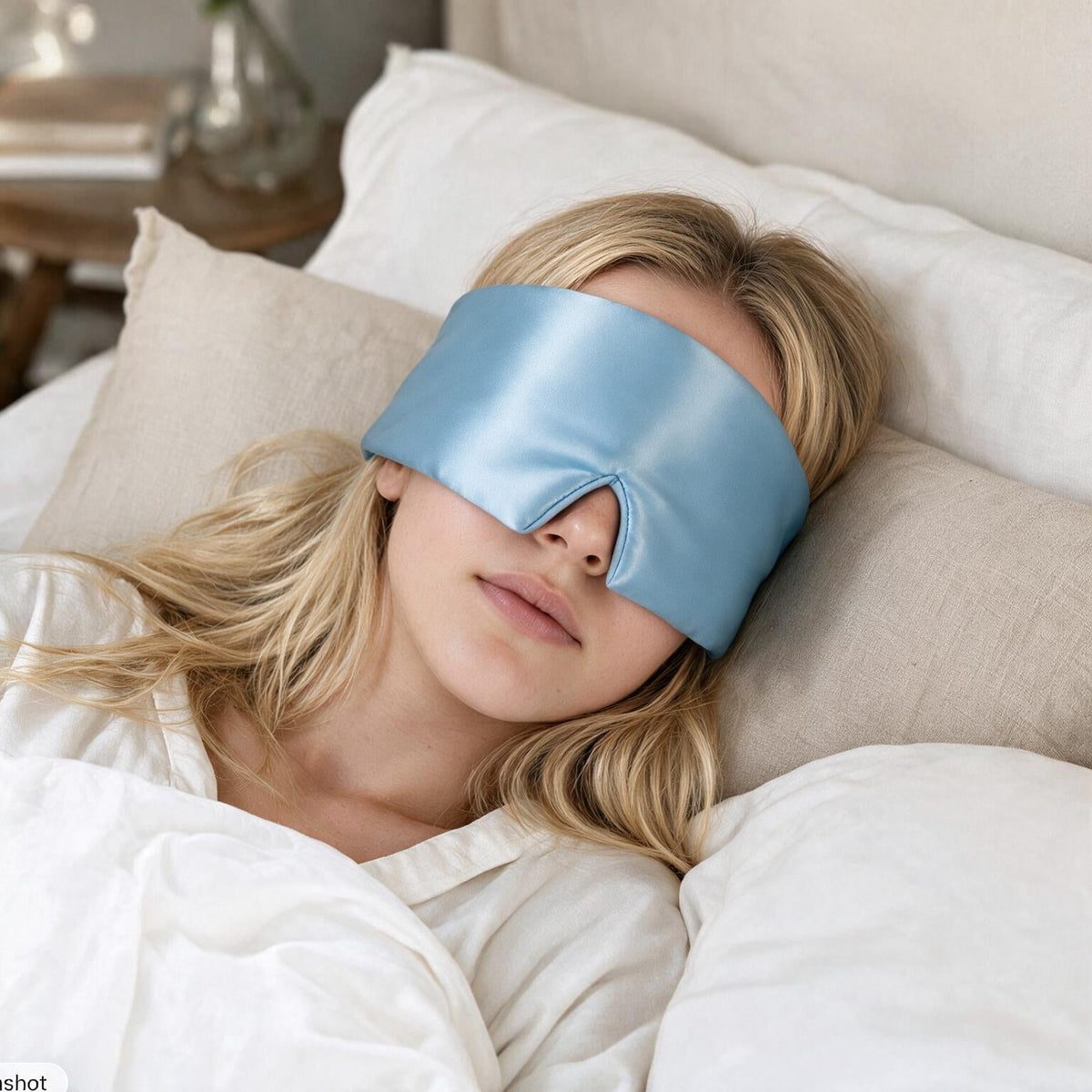 Woman asleep on her side wearing the Lunia Reve mulberry silk sleep mask in Sky Blue on white cotton bedding