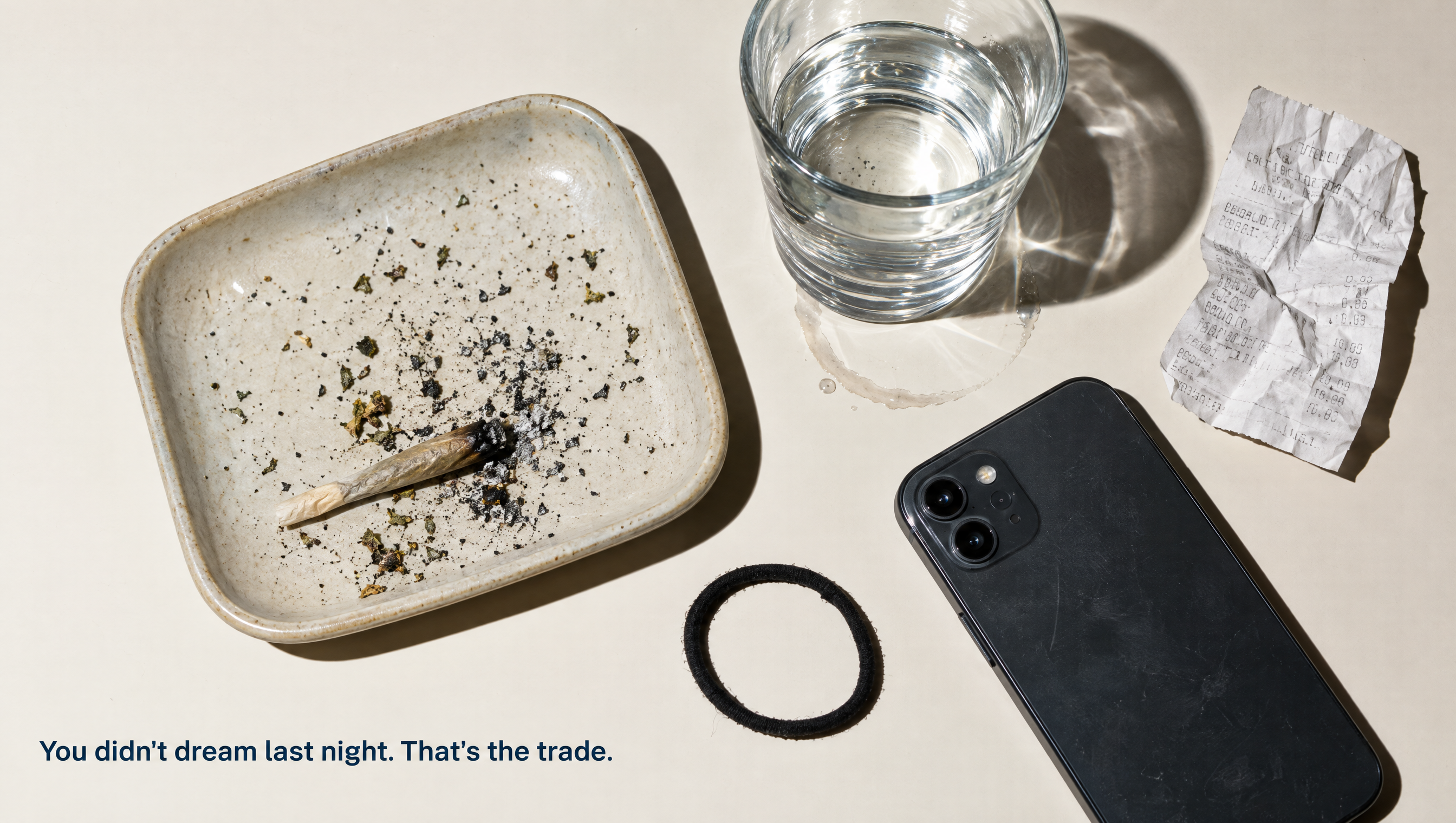 Overhead view of a nightstand at night: a ceramic tray holding a stubbed-out joint and loose ash, a phone lying face down, a glass of water and a hair tie on a pale surface.