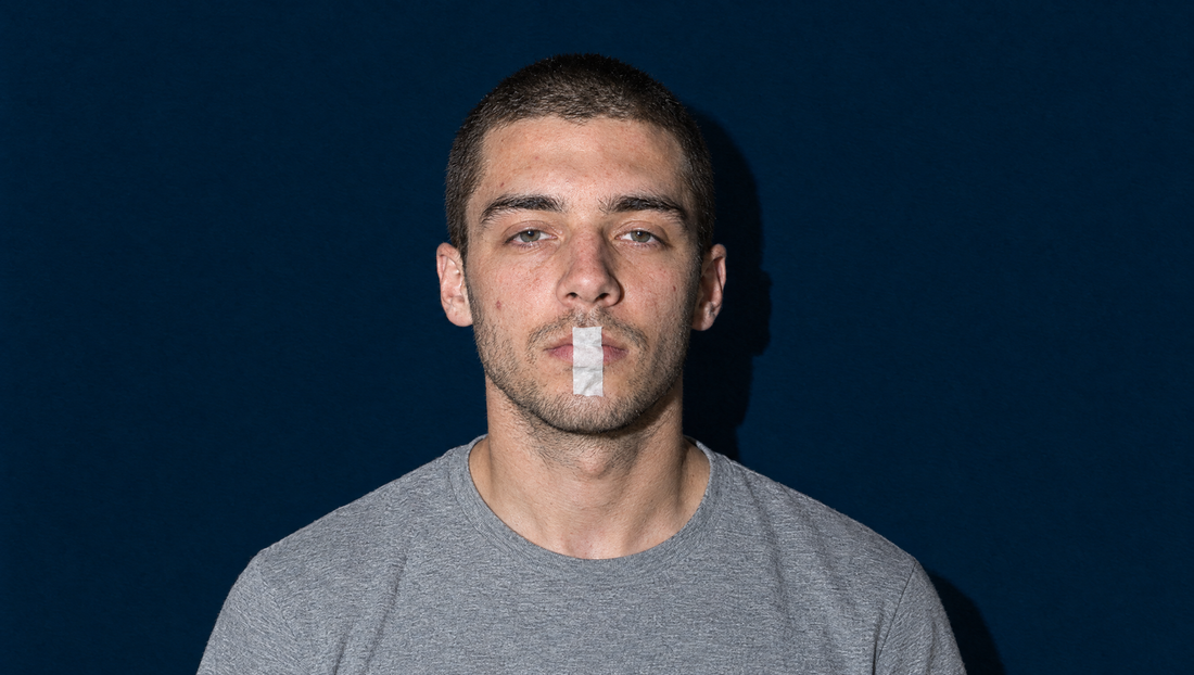 Hard-flash portrait of a young man with a buzz cut in a grey t-shirt against a flat navy backdrop, a single vertical strip of white paper tape across his closed lips