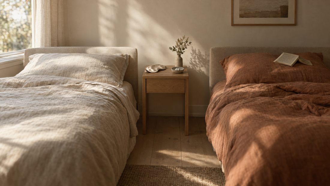 Two separate single beds in a bright bedroom, one with an oat linen duvet and one with a clay duvet, with an oak nightstand between them holding a folded cream eye mask and a small dish of white foam earplugs