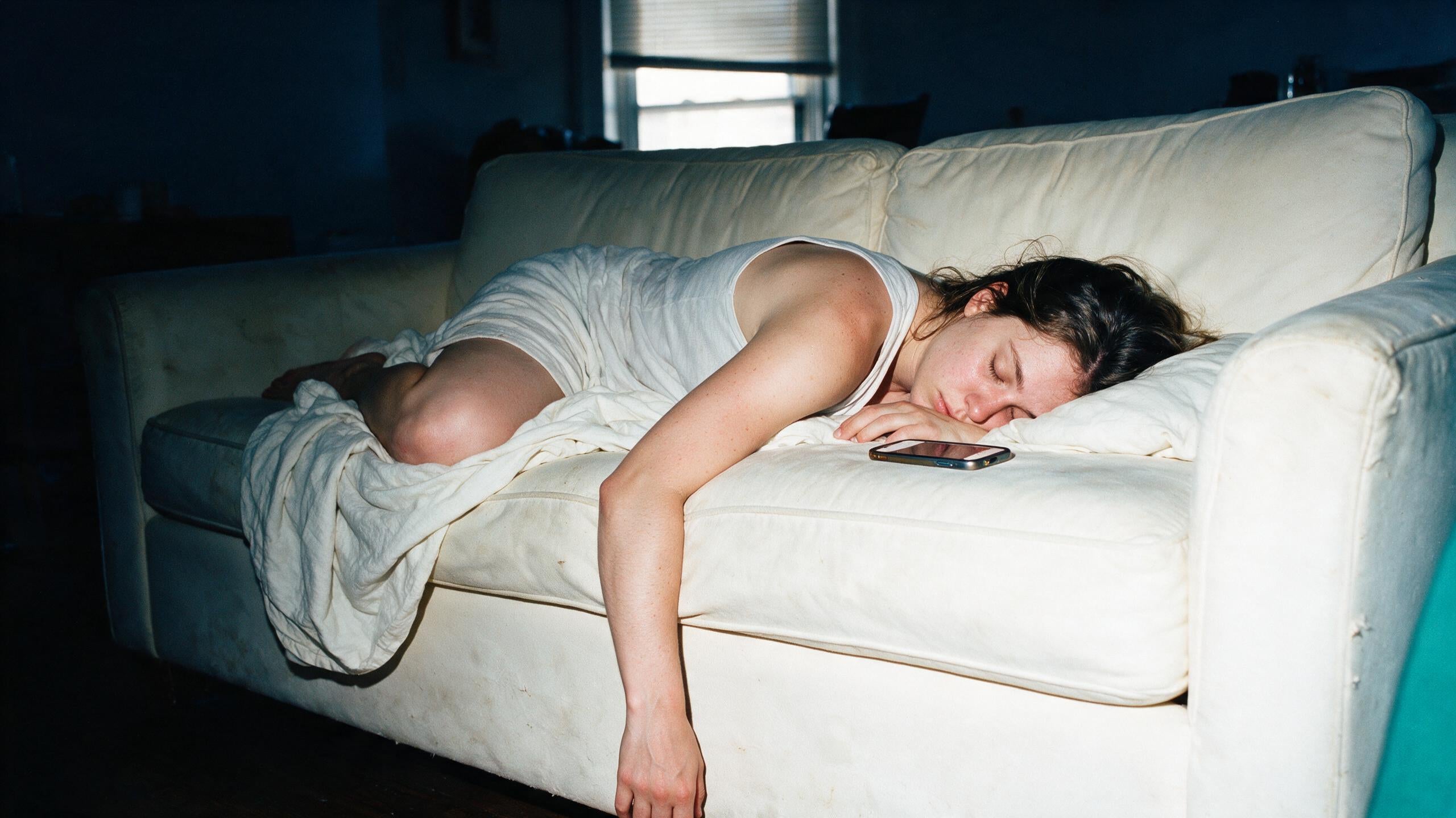 A young woman asleep face down on a worn ivory couch in a dim apartment living room, her phone lying face up on the cushion beside her hand, shot with direct camera flash