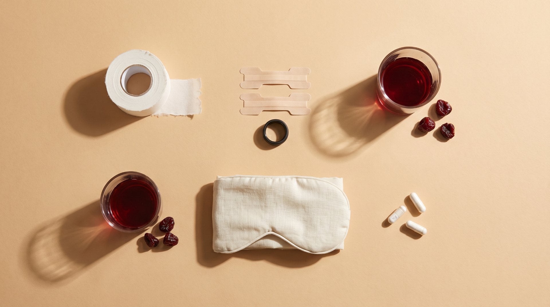 Overhead flat lay on sand-colored paper of the sleepmaxxing toolkit: a roll of white tape, nasal strips, a black smart ring, two glasses of tart cherry juice, a cream eye mask and loose white capsules